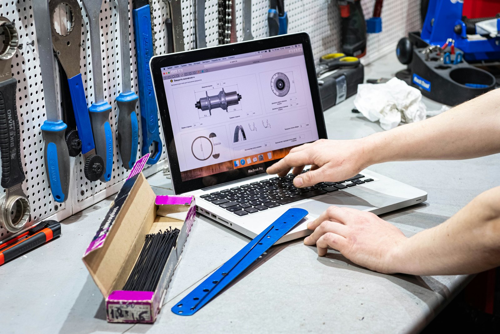 Hands typing on a laptop on a workshop bench, with hand tools hanging on a pegboard behind it