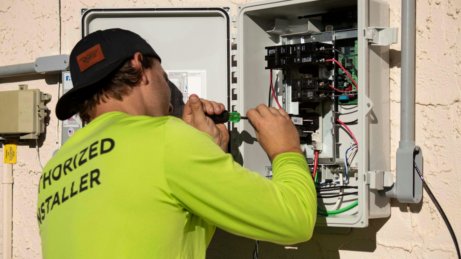 An electrician in a bright yellow shirt using a screwdriver on an open electrical panel mounted on a house wall