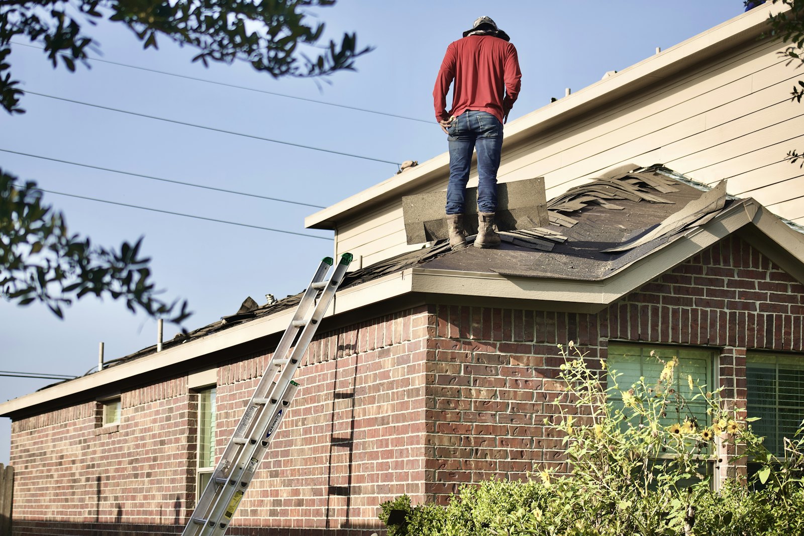 A roofer in a red hoodie standing on a half stripped roof of a brick house, with a ladder against the gutter