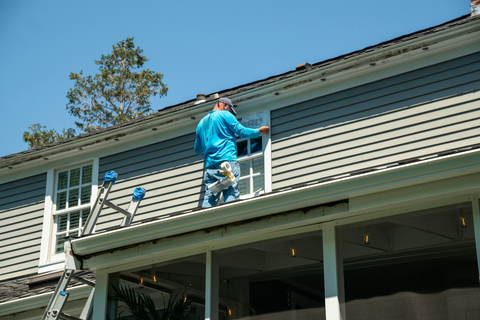 An installer in a blue shirt taping around a window on a house with gray lap siding, a ladder leaning on the porch roof