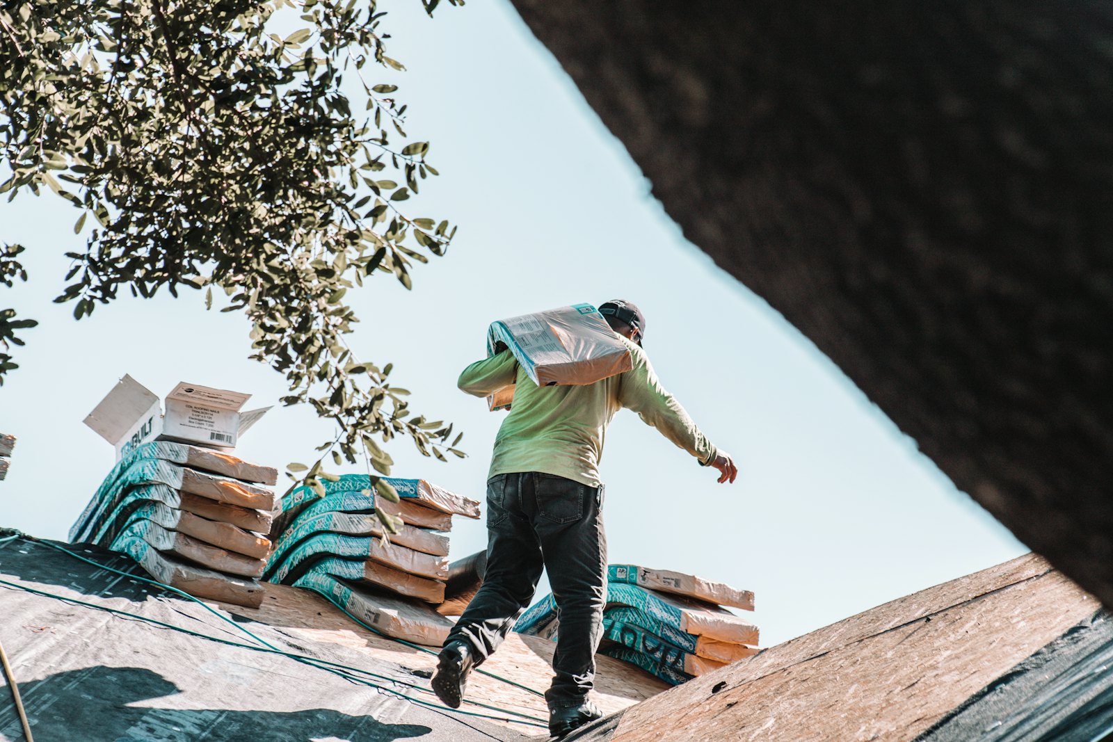 A roofer carrying a bundle of shingles across a roof deck, with more bundles stacked beside him